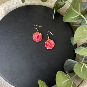 May include: A pair of gold hoop earrings with pink and red marbled round dangles. The earrings are on a black surface.