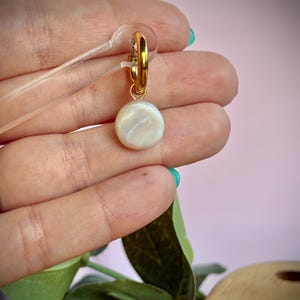 May include: A gold-colored hoop earring with a round, pearlescent charm. The charm has a swirling pattern and a subtle sheen. The earring is held up by a clear plastic earring display card. The background is a soft pink.