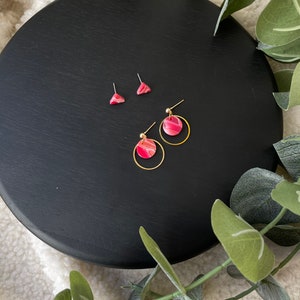 May include: A set of three pink and gold earrings. Two earrings are small triangles and the third earring is a circle with a pink and white swirl design.