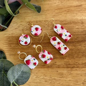 May include: A collection of handmade earrings featuring a strawberry theme. The earrings have a pink and white checkered pattern, adorned with red strawberries and small white flowers. The earrings are in various shapes, including circles and rectangles, with gold-colored hooks.