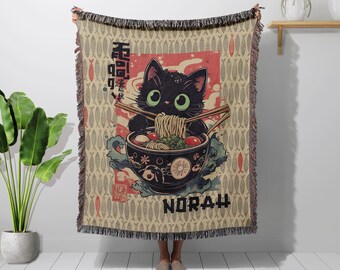 May include: A beige throw blanket featuring a black cat with green eyes, holding chopsticks and eating ramen from a decorative bowl. The design includes Japanese text, red accents, and the name "NORAH" at the bottom. The blanket has a fringe.