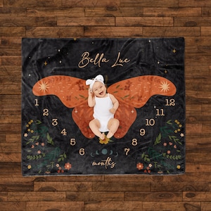 May include: A black baby blanket with a large orange butterfly design. The butterfly has floral details and a star on each wing. The blanket has numbers 1-12 around the butterfly and the text "Bella Lue" and "months" at the bottom.