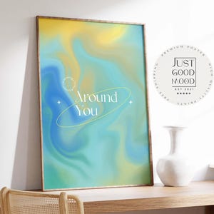 Puede incluir: Un cuadro enmarcado con un diseño abstracto colorido. La obra presenta una mezcla de tonos amarillos, azules y verdes con las palabras "Around You". Un jarrón blanco se encuentra sobre una superficie de madera.