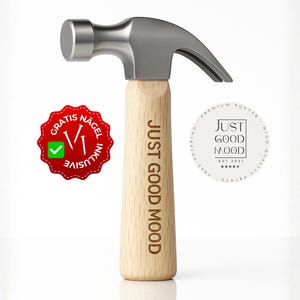 Puede incluir: Un martillo con cabeza plateada y mango de madera clara. El mango está grabado con las palabras "JUST GOOD MOOD". Una pegatina roja con las palabras "GRATIS NAGEL INKLUSIVE" y una marca de verificación está a la izquierda. Un logotipo circular blanco está a la derecha.