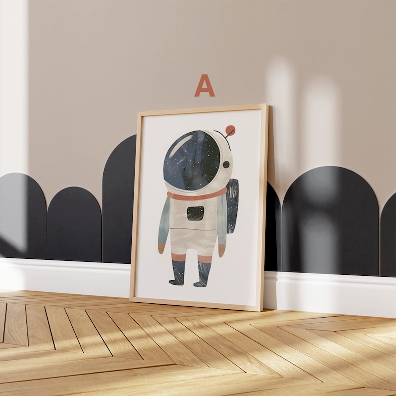 May include: A framed print of a cartoon astronaut with a starry helmet, wearing a white spacesuit with a red stripe and a blue and white pattern. The print has a white background and the letter "A" is printed in the top center of the frame.