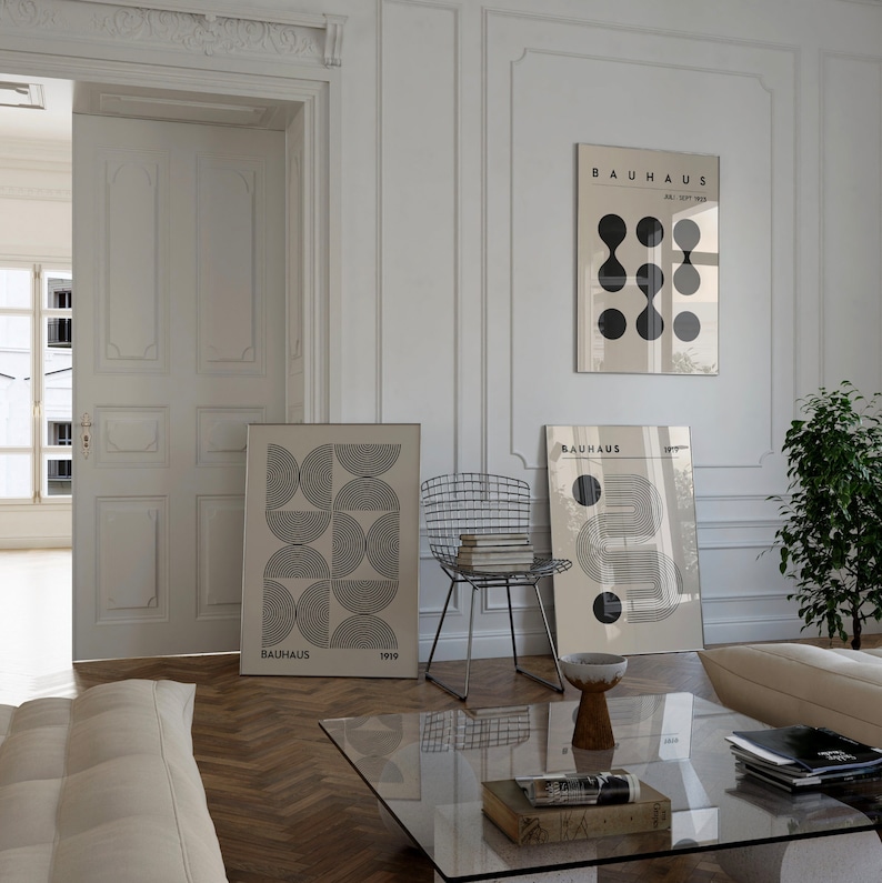 Puede incluir: Tres impresiones de arte geom&eacute;tricas minimalistas en blanco y negro con el texto "Bauhaus" y los a&ntilde;os "1919" y "Abril-Sept 1923" en una pared blanca en una sala de estar moderna con una mesa de caf&eacute; de vidrio, un sof&aacute; blanco y una silla de metal.