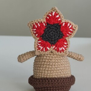 May include: A crochet amigurumi figure featuring a tan body and a dark brown base. The head is a star-shaped flower with a dark gray center, red petals, and white stitched teeth along the edges. Two small tan arms extend from the sides of the body.