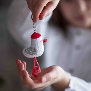 Può includere: Un portachiavi a forma di gabbiano all'uncinetto con un cappello rosso e le gambe. Il gabbiano è bianco con le ali grigie e ha un becco rosa.