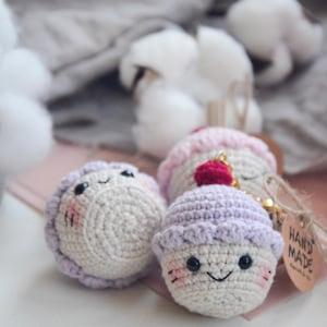 May include: Three crocheted cupcake keychains with smiling faces. The cupcakes are white with purple and pink frosting and red cherry accents. The tag reads "HAND MADE".