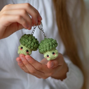 Puede incluir: Dos llaveros de brócoli tejidos a crochet con caras lindas, verde y verde claro, colgando de una cadena plateada.