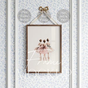 May include: A framed art print featuring three ballerinas in pink and green tutus. The artwork is displayed in a brown wooden frame, hanging from a gold bow. The background is a floral wallpaper with white molding.