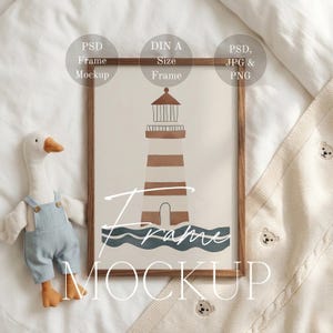 Puede incluir: Un marco con una ilustración de faro en marrón y blanco, sobre un fondo crema. El marco de madera rodea la obra de arte. También se ven un juguete de peluche de ganso y una manta crema con bordados de osos. El texto "Frame Mockup" está en la impresión.