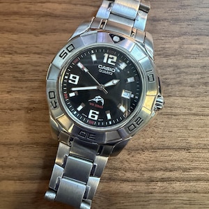 May include: A silver-tone stainless steel Casio Quartz analog watch with a black dial, featuring a rotating bezel with engraved numbers, a date window at the three o'clock position, luminous hands, and a marlin logo above the WR 20BAR text.