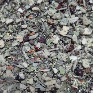 May include: Close-up of a mix of dried herbs and berries. The blend features various shades of green leaves, interspersed with dark red and brown berries and small pieces of orange. The texture is rough and natural, suggesting a herbal tea or spice mix.