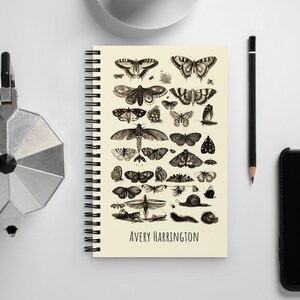 May include: A spiral bound notebook with a cream cover featuring a vintage illustration of various butterflies and moths. The notebook has the name "Avery Harrington" printed at the bottom.
