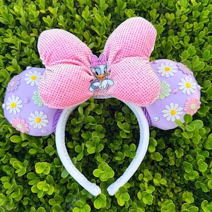 May include: A headband featuring two light purple ears decorated with white, yellow, and pink daisy appliques and small rhinestones. A large, textured pink bow sits between the ears, centered with a small Daisy Duck character emblem.