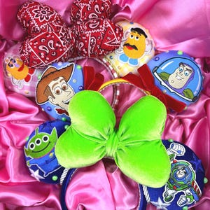 May include: Two headbands featuring embroidered character patches from Toy Story, including Woody, Buzz Lightyear, Mr. Potato Head, and an alien. One headband has a large red paisley bow, and the other has a bright lime green velvet bow.