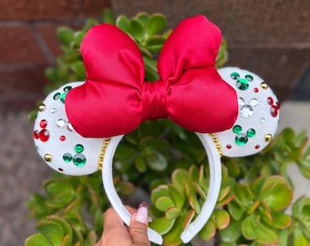 May include: A white headband with two white mouse ears decorated with green, red, and gold accents. A large red satin bow sits atop the headband. The headband is surrounded by green plants.