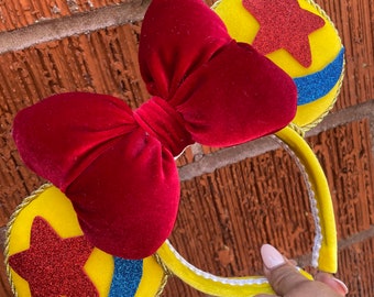 May include: Yellow mouse ear headband with a large red velvet bow. The ears are yellow with a red glitter star and a blue glitter stripe. The headband is held by a hand.