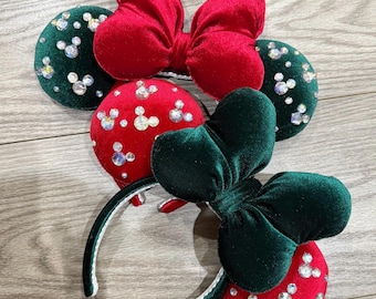 May include: Two sets of headband ears featuring plush velvet fabric. One headband has dark green ears with clear crystal embellishments and a large red velvet bow. The other headband has bright red ears with clear crystal accents and a large dark green velvet bow.