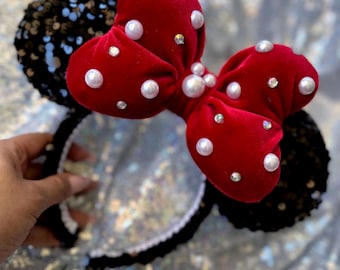 Op de afbeelding: Een zwarte haarband met pailletten oren en een rode fluwelen strik. De strik is versierd met witte parels en kleine heldere strass-steentjes. Een leuk accessoire.
