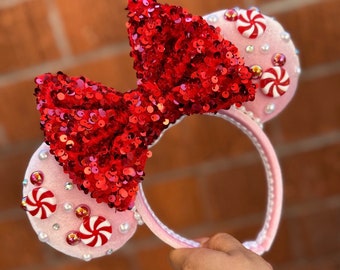 May include: A pink headband with Minnie Mouse ears, adorned with red sequined bow and candy-themed embellishments. The ears are light pink with white and red peppermint candies and pearl accents. A festive accessory.