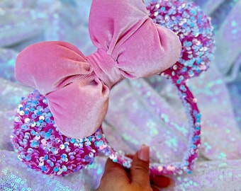 Peut inclure: Un serre-tête avec un nœud en velours rose et des paillettes roses irisées. Le serre-tête est sur un fond blanc iridescent.