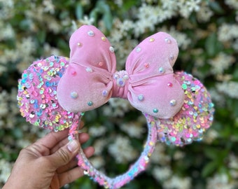 May include: Pink velvet bow headband with iridescent sequins and pearl-like embellishments. The headband is a popular accessory for Disney theme parks.