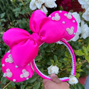 May include: Pink satin Minnie Mouse ears headband with a large bow and white pearl and rhinestone embellishments.