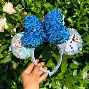 May include: A headband featuring a large, bright blue sequined bow at the center. The two circular ears are covered in a light blue fabric; one ear displays an illustration of a blonde character, while the other features a white snowman character.