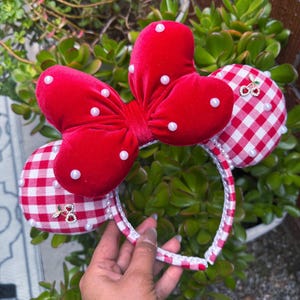 May include: A red velvet bow headband with white pearl accents and red and white gingham ears. The headband features a red bow with white polka dots and small pearl embellishments. The ears are decorated with a cherry charm.