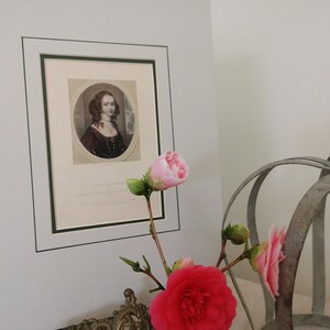 May include: A framed portrait of a woman with dark hair and a white lace collar, wearing a dark dress. The portrait is in a green frame and is hanging on a white wall. There are pink flowers in the foreground.