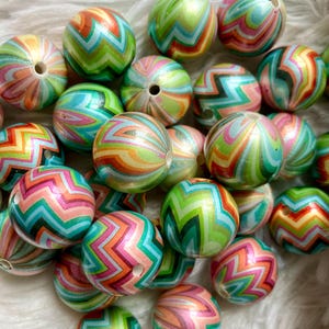 May include: A selection of round beads with a chevron pattern in green, orange, pink, and blue. Each bead has a hole for threading. The beads are scattered on a white, fluffy surface. The beads are approximately 2 cm in diameter.