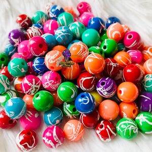May include: A pile of vibrant, spherical beads in shades of blue, green, pink, orange, red, purple, and yellow. Each bead features a white, swirled pattern. The beads are arranged on a white, fluffy background.