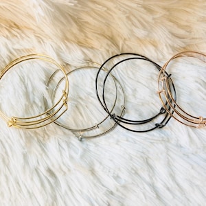 May include: Four expandable bangle bracelets in gold, silver, and black metal. The bracelets are stacked on a white fluffy surface.