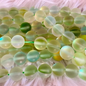 May include: Strands of round, frosted glass beads in shades of green, yellow, and white. The beads are strung on green thread, creating a gradient effect. The beads have a soft, iridescent sheen.