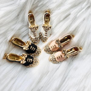 May include: Four gold-tone shoe charms with rhinestones. Two are white with black trim and bows, and two are pink with gold trim and bows.