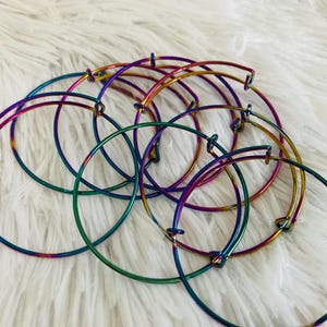 May include: A collection of iridescent metal bangle bracelets in shades of green, blue, purple, and gold. The bracelets are arranged on a soft, white, textured surface. The bracelets are adjustable.