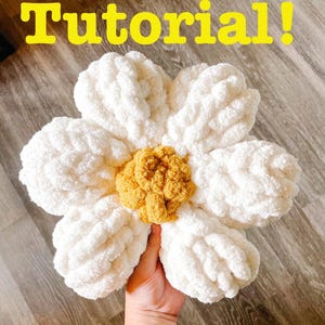 Op de afbeelding: Een grote, handgemaakte, witte en gele gehaakte bloem. De bloem heeft een donzige textuur en wordt in een hand gehouden. Het woord "Tutorial!" staat in het geel bovenaan de afbeelding.