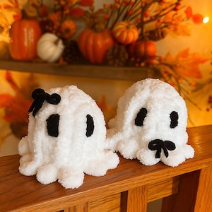 May include: Two handmade white plush ghost figures with black eyes and bows. The ghosts are sitting on a wooden surface, with a fall-themed background of pumpkins and foliage.