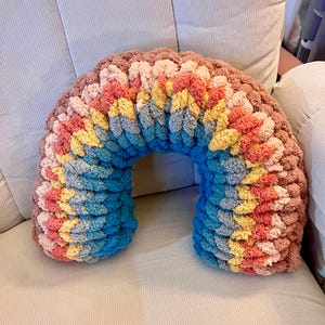 May include: A rainbow-colored crocheted pillow in the shape of an arch. The pillow is made with chunky yarn and has a soft, fluffy texture.