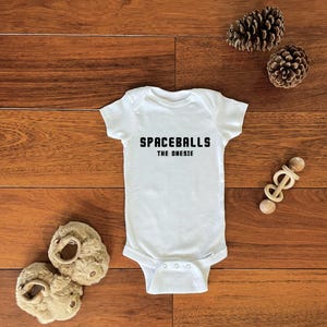 May include: White baby onesie with the text "SPACEBALLS THE ONESIE" in black. The onesie is laid on a wooden surface with a pair of beige baby shoes, two pinecones, and a wooden toy.