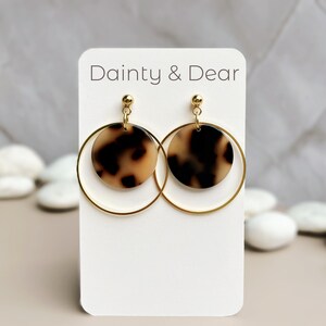 May include: Gold hoop earrings with a brown and cream tortoiseshell patterned circle pendant. The earrings are displayed on a white card with the text "Dainty & Dear".