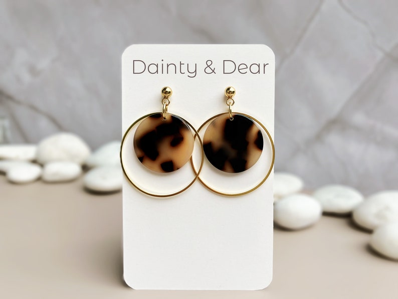 May include: Gold hoop earrings with a brown and cream tortoiseshell patterned circle pendant. The earrings are displayed on a white card with the text "Dainty & Dear".