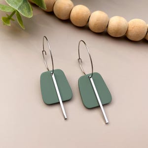May include: A pair of sage green earrings with a silver bar pendant. The earrings have a geometric shape and are suspended from silver hoops. The earrings are displayed on a neutral background with wooden beads and greenery.