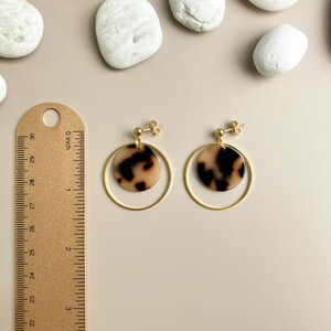 May include: Gold hoop earrings with a brown and white patterned circle pendant. The earrings are shown next to a ruler.