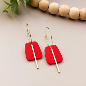 May include: A pair of red and gold earrings. Each earring features a red, rectangular shape with rounded corners, suspended from a gold hoop. A gold bar hangs vertically from the center of each red shape.