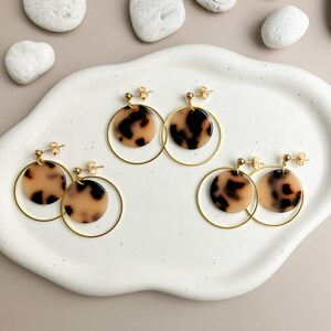 May include: Four pairs of gold hoop earrings with a brown and cream patterned circle pendant. The earrings are displayed on a white ceramic plate.