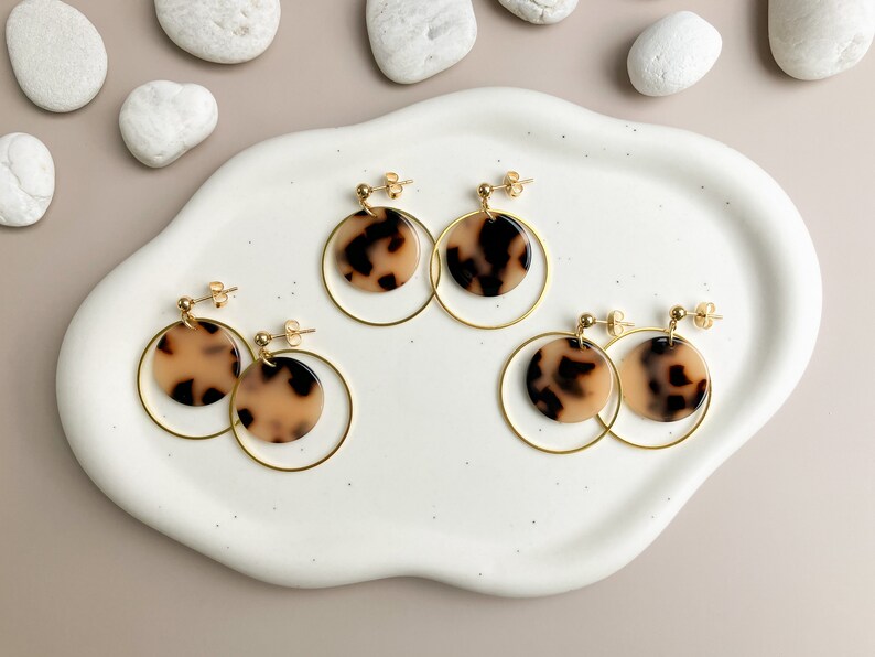May include: Four pairs of gold hoop earrings with a brown and cream patterned circle pendant. The earrings are displayed on a white ceramic plate.
