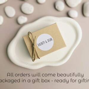 May include: A small brown box with a white circle sticker that says "DAINTY & DEAR" on it. The box is tied with a gold ribbon and is sitting on a white ceramic dish.
